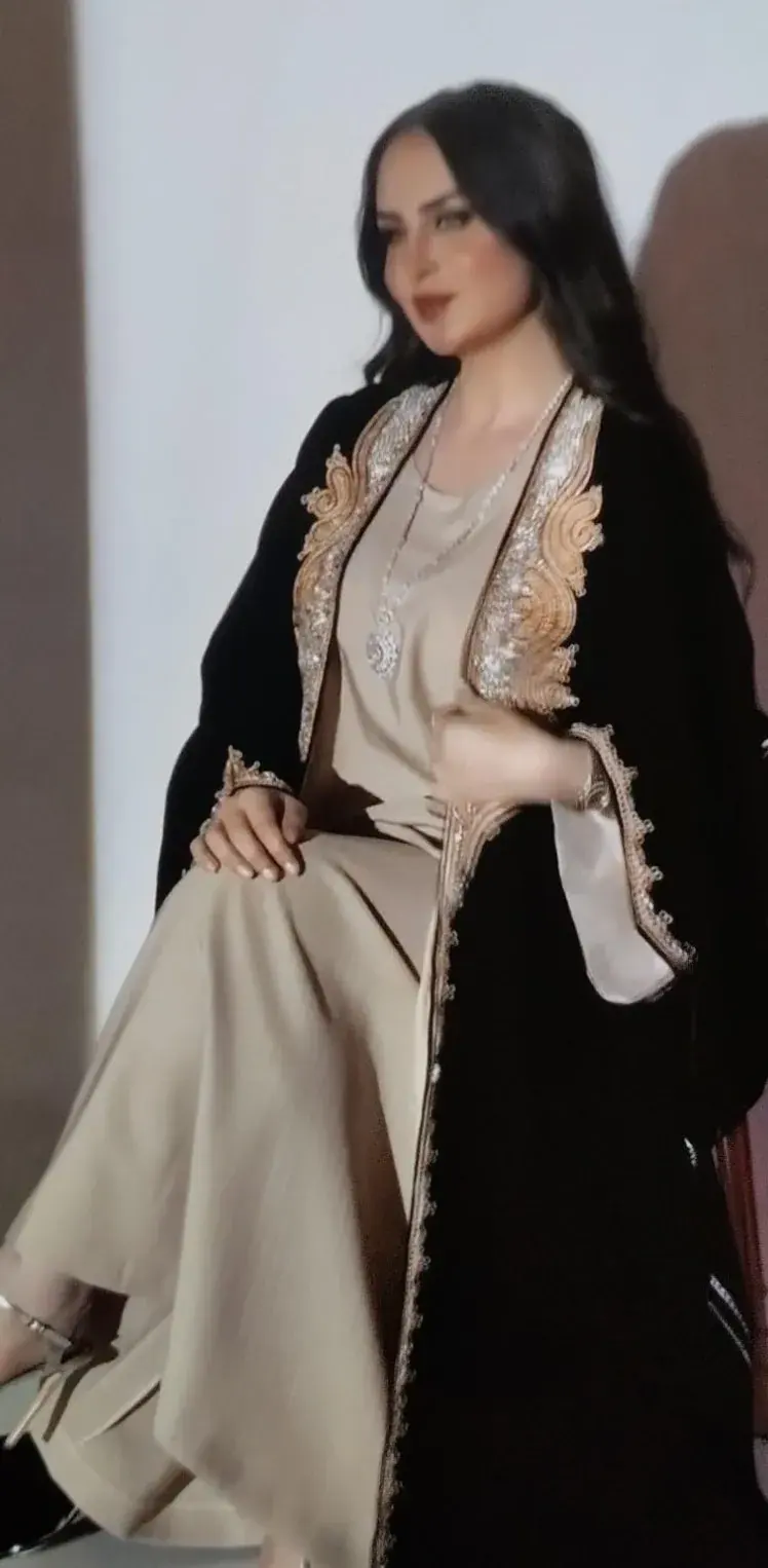 Model seated in a black velvet robe with gold embroidery over a sand dress